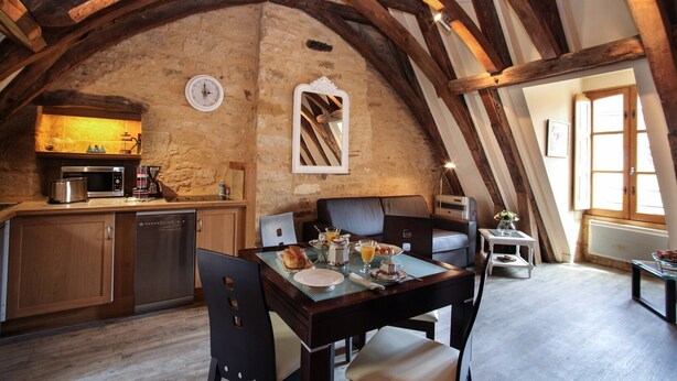 Cocina rústica con vigas de madera, pared de piedra y zona de comedor con una mesa dispuesta para el desayuno.