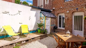 A courtyard with a wooden table and chairs, a small garden area with potted plants, and a brick building with a door and windows.