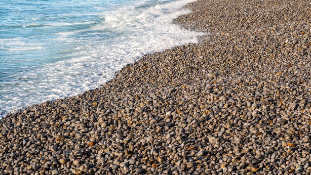 A pebbled beach with a large rock formation and a clear blue sky.