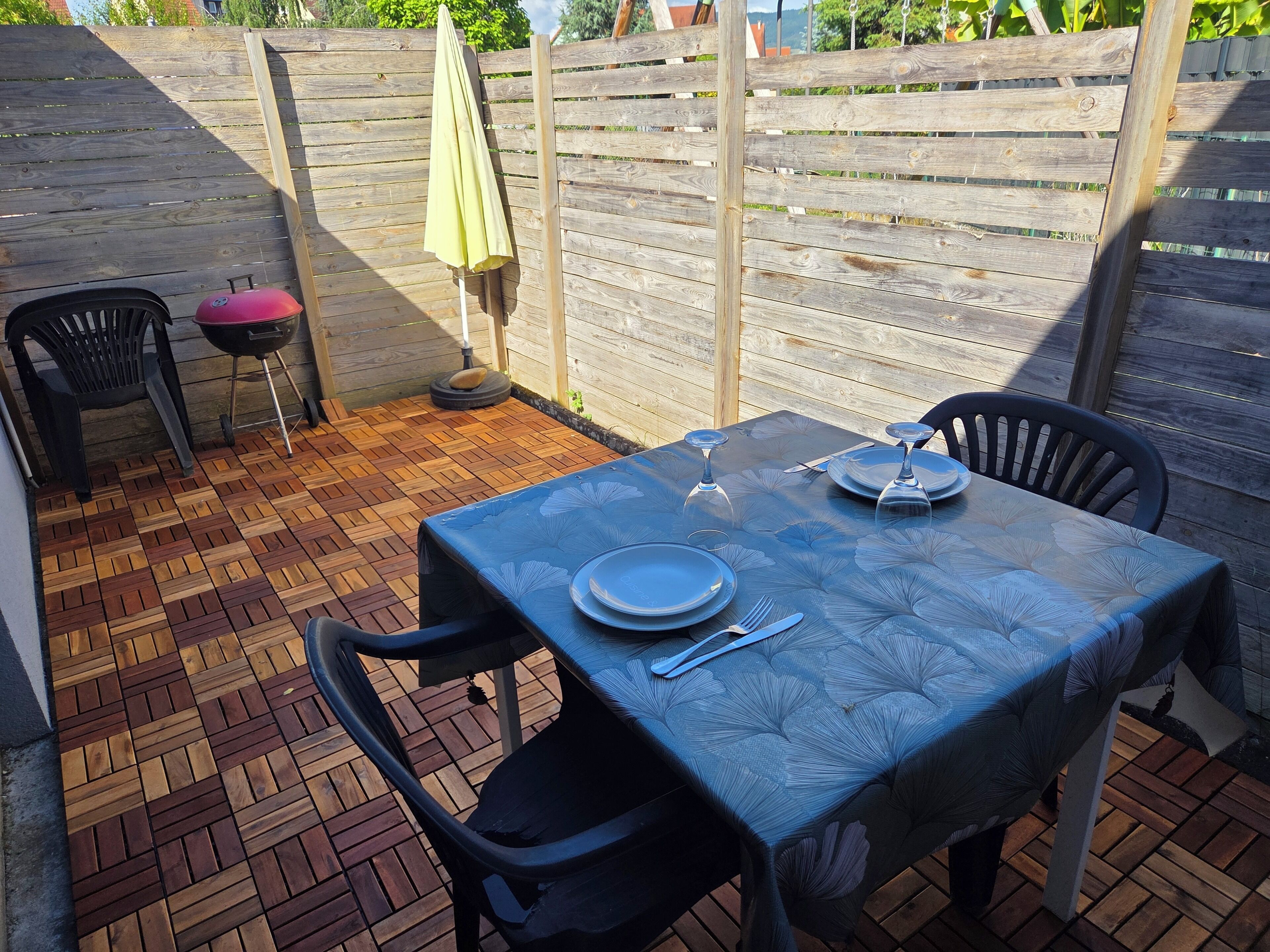 Un patio con una mesa dispuesta para dos personas, dos sillas, una parrilla y una valla de madera al fondo.
