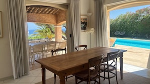 A dining area with a wooden table and chairs, large windows with curtains, and a view of a swimming pool and tropical landscape. - Superb villa with panoramic sea view Sainte Maxime (Sainte-Maxime)