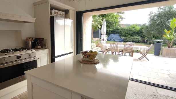 Une cuisine avec des meubles blancs, une cuisinière à gaz et un réfrigérateur. Il y a également un coin repas avec une table et des chaises à l’extérieur.