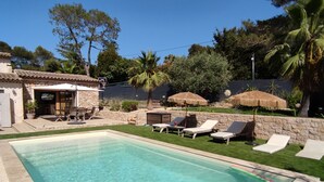 Une piscine rectangulaire à l’eau claire, entourée d’une terrasse en pierre et d’une pelouse aménagée avec des chaises longues et des parasols.