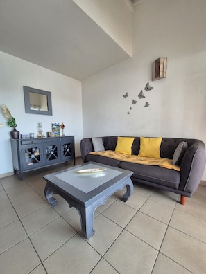 Uma sala de estar com um sofá cinzento, uma mesa de centro de vidro, um armário de madeira escura e um espelho fixado na parede.