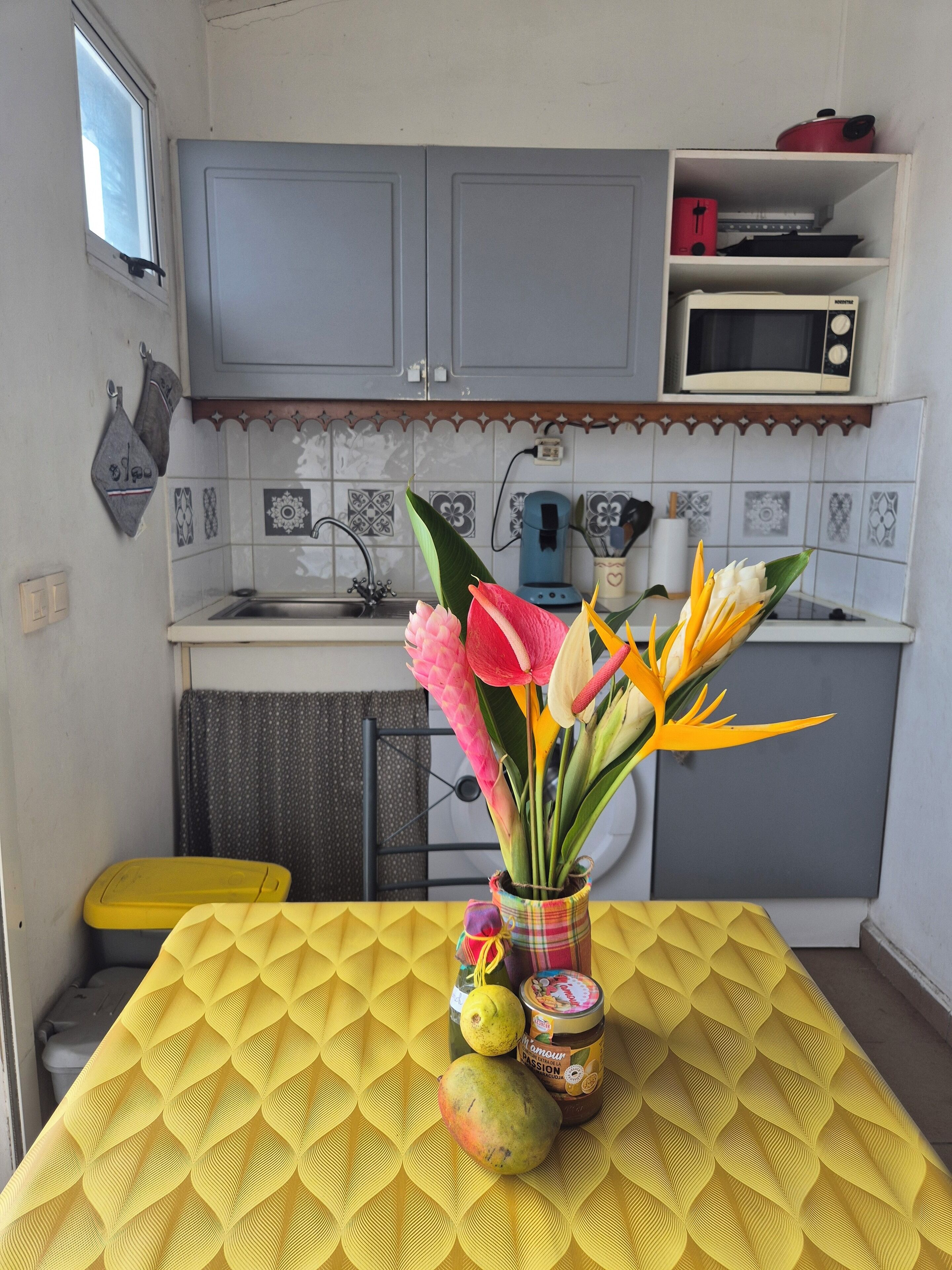 Una cocina con gabinetes grises, un microondas y un lavamanos. Hay una mesa amarilla con mantel estampado, un florero con flores y un tarro de mermelada.