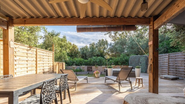 Een terras buiten met een houten tafel en stoelen, een barbecue en een overdekte zithoek.
