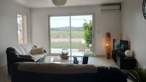 Uma sala de estar com um sofá, uma mesa de centro de vidro, uma televisão e vista para uma paisagem verde através de uma porta de correr.