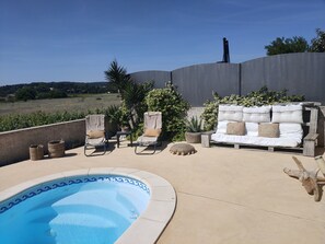 Zona de piscina con un sofá blanco y dos sillones de descanso, rodeada de plantas en macetas y vista a un campo.