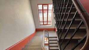 A spiral staircase with a wooden handrail and metal balusters, leading to a landing with a red-painted baseboard and a window with a view of buildings outside.