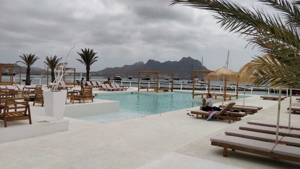 A poolside area with lounge chairs and a pool, surrounded by palm trees and mountains in the background. - Apartment for rent. on the island of Sao Vicenter Mindelo near the beach (Mindello)