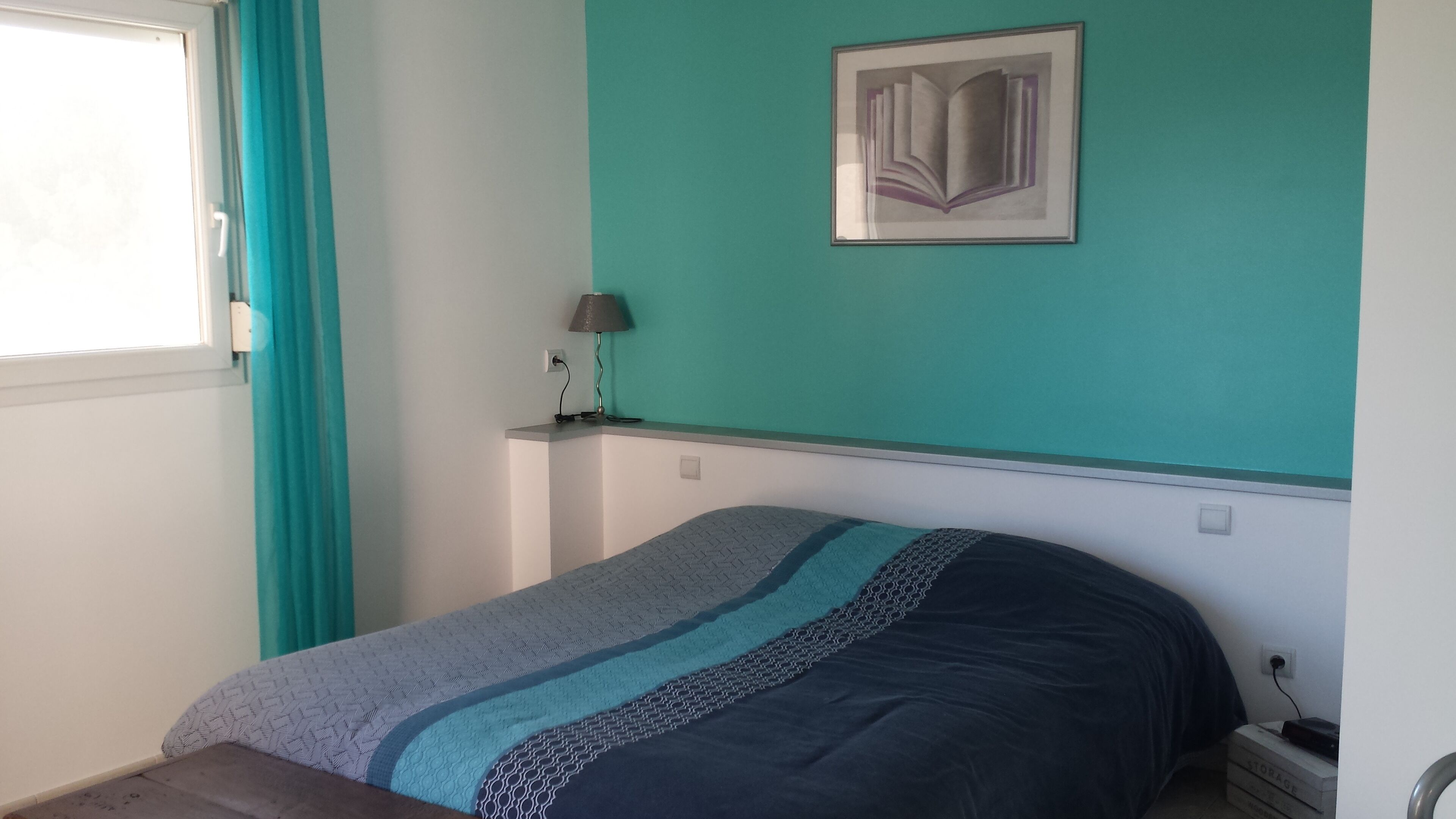 Une chambre à coucher avec un lit, une lampe fixée au mur et un tableau encadré.