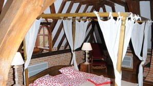 Une chambre à coucher avec un lit à baldaquin à quatre poteaux, des poutres en bois, un fauteuil rouge et une fenêtre donnant sur un autre fauteuil rouge à l’extérieur.