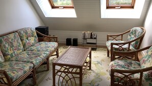 Une pièce mansardée chaleureuse avec un plafond en pente, deux fenêtres de toit, un canapé à motifs floraux, des fauteuils en osier, une petite table et une bibliothèque.