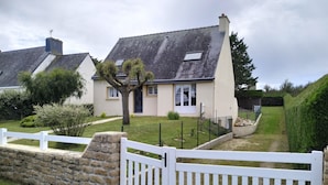 Une maison à deux étages, avec un toit en pente, un mur en pierre et une clôture blanche faite de piquets.