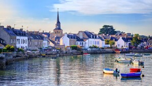 Une ville côtière dotée de bâtiments historiques, d’un clocher d’église imposant et d’un port tranquille où sont amarrés de petits bateaux.
