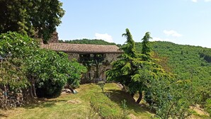 Un bâtiment en pierre, surmonté d’un toit en tuiles, entouré d’une végétation luxuriante et d’arbres.