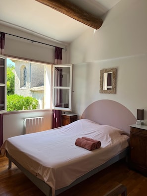 A bedroom with a bed, a window with curtains, a radiator, a wooden headboard, a bedside table, and a mirror.