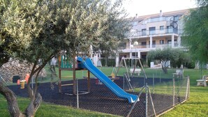 Un parque infantil con un tobogán azul y columpios, rodeado por una cerca y árboles.