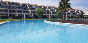 Una piscina con bordeado de azulejos, rodeada de un área ajardinada con palmeras y un edificio al fondo.