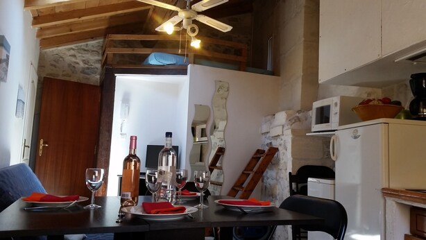 Uma sala de jantar com uma mesa posta para quatro pessoas, um ventilador de teto e uma cozinha equipada.
