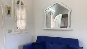 A room with a blue sofa, a white side table with a bowl, a decorative mirror, and a window with a curtain. - Dar Amandine Djerba midoun (Midoun Djerba)