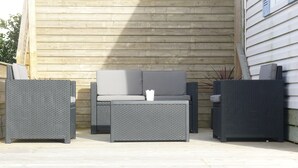 Een terras buiten met rieten meubels, waaronder een bank en twee fauteuils, tegen een houten wand.