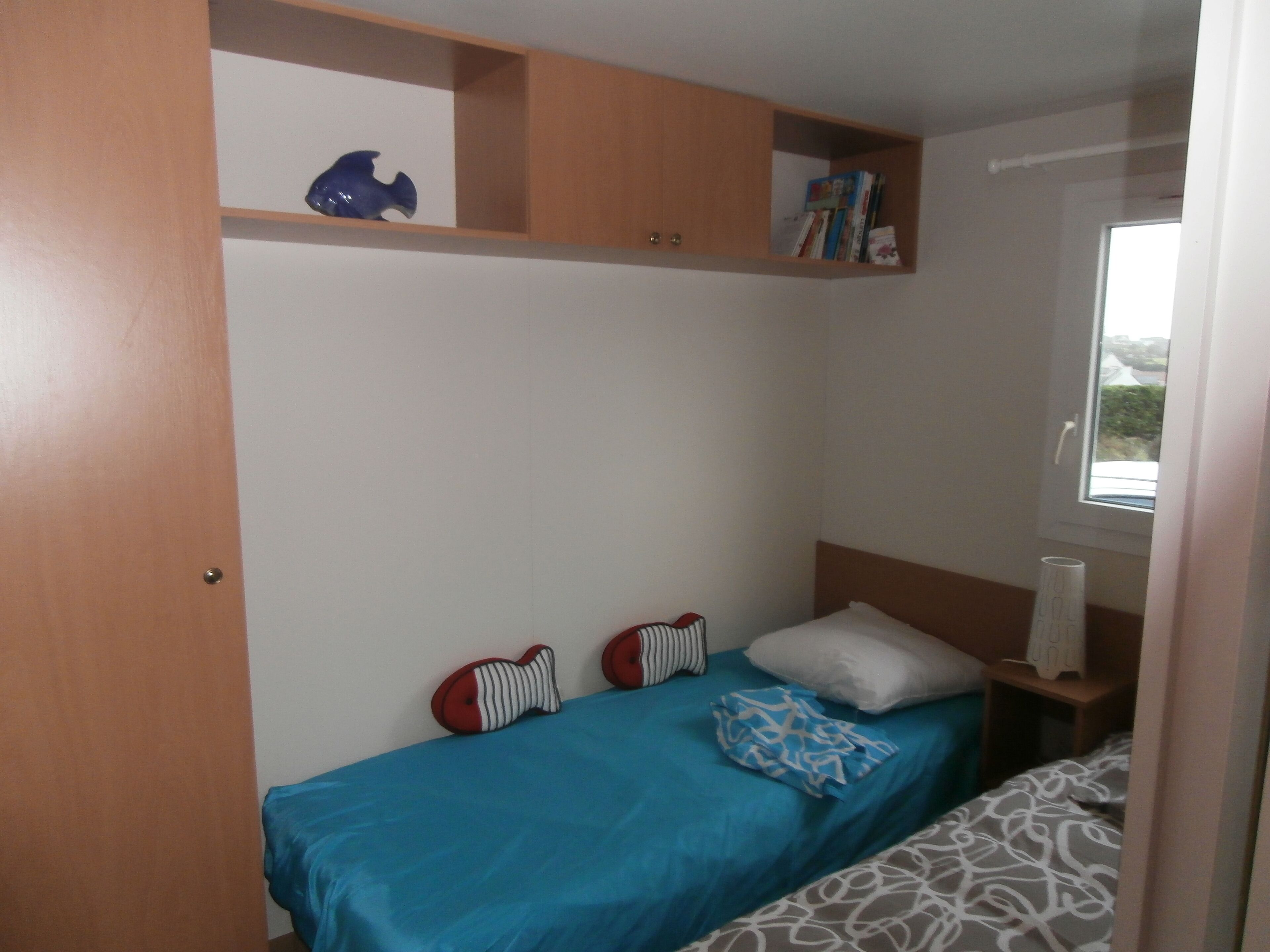 Une chambre avec un lit simple, une table de chevet, une étagère avec des livres et un objet décoratif, et une fenêtre donnant sur l’extérieur.