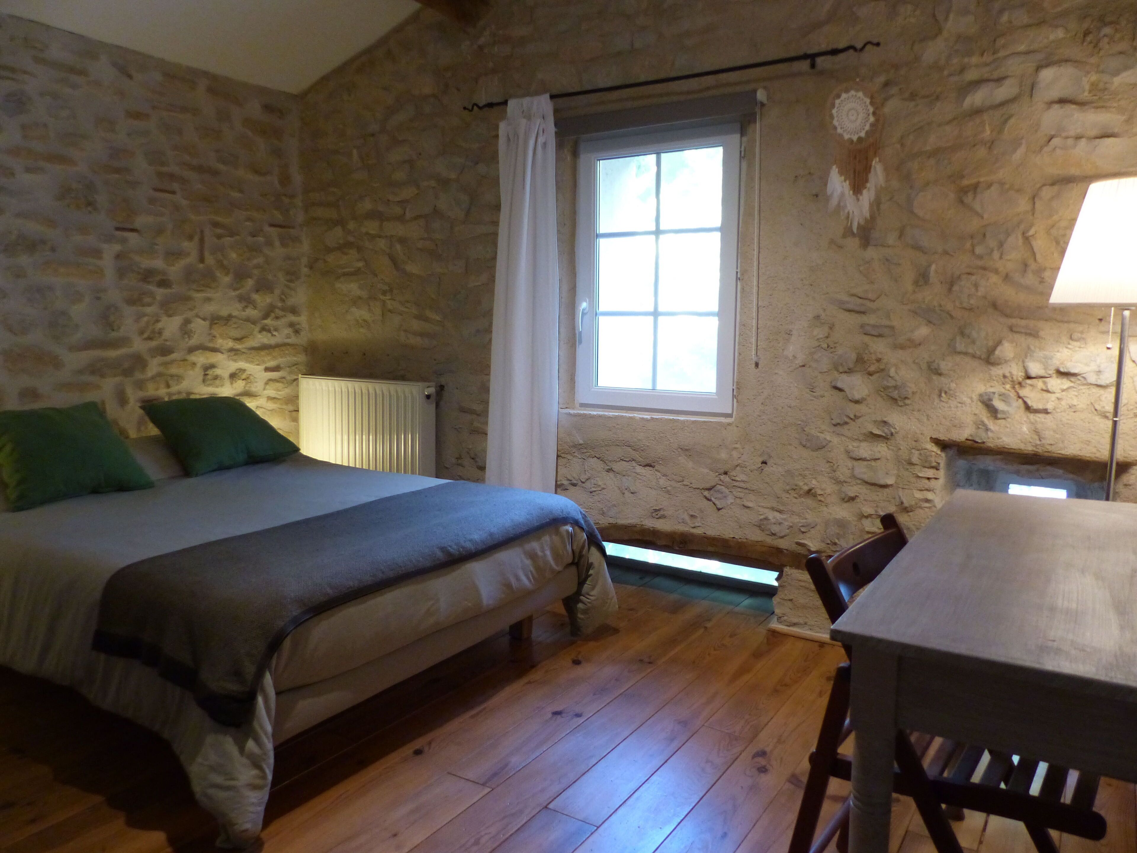 A room with a stone wall, a bed with green pillows, a window with white curtains, a radiator, and a wooden table.