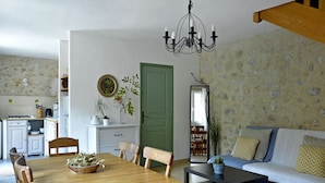 Een zithoek met een houten tafel en stoelen, een bijzettafeltje en een bank met kussens. Er is een groene deur, een spiegel en een kroonluchter.
