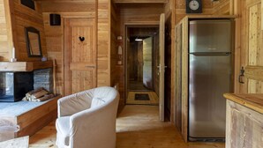 A wooden cabin interior with a fireplace, a white armchair, a stainless steel refrigerator, and a wooden ceiling.