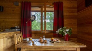 A wooden cabin interior with a dining table set for breakfast, a window with curtains, and a view of greenery outside.