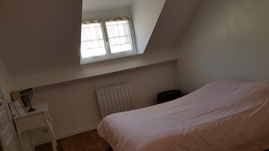 A bedroom with a sloped ceiling, a bed with a patterned cover, a white nightstand, a radiator, and a window with curtains. - In the heart of the city center (Blois)