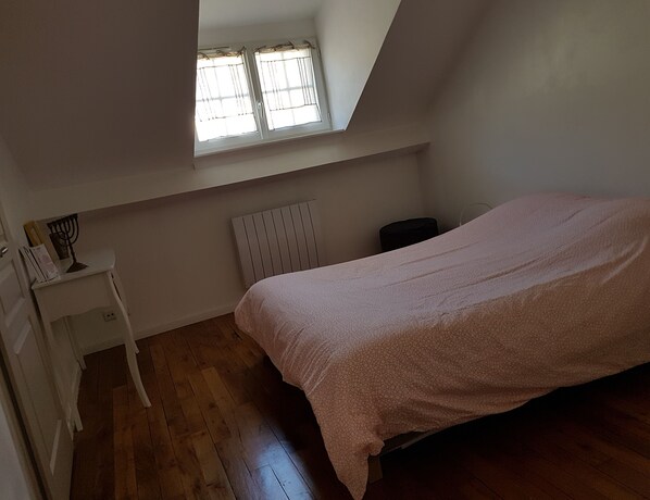Une chambre avec un plafond en pente, un lit recouvert d’une housse à motifs, une table de chevet blanche, un radiateur et une fenêtre avec des rideaux.