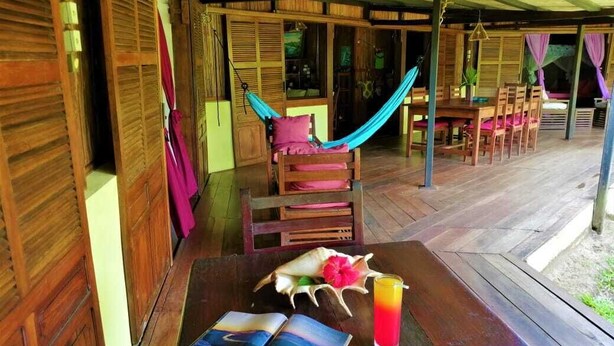 Een houten terras met een hangmat, een tafel met een drankje en een tijdschrift, en een houten stoel.