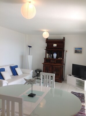 Una sala de estar con un sofá blanco, una mesa de centro de vidrio, un aparador de madera, un televisor y un ventilador.