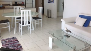 Cocina moderna con armarios blancos, microondas y refrigerador. Hay un comedor con mesa de vidrio y sillas blancas. En la sala de estar, una mesa de centro de vidrio y un sofá blanco con un cojín azul son visibles.