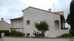 Una casa de dos pisos con techo de tejas, un árbol pequeño en la parte delantera y un muro bajo con una reja.