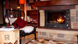 A cozy living room with a fireplace, a checkered floor, a wooden side table, a chair with a white fur throw, and a bookshelf.