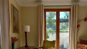 A room with a wooden chair, a table with a lamp, a bed, and a view through a glass door to an outdoor area with plants.