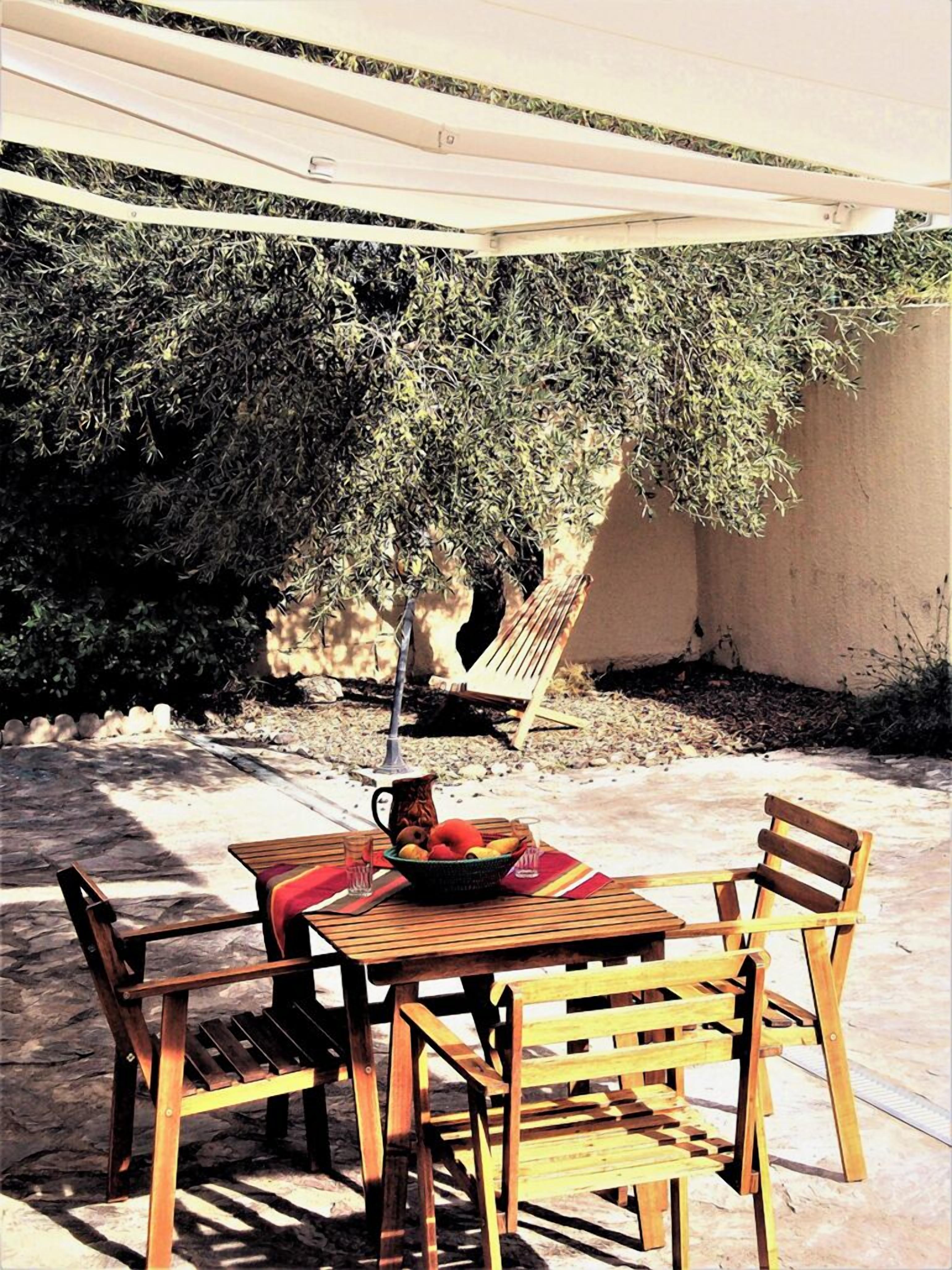 An outdoor patio with a wooden table and chairs, a pitcher, and a bowl of fruit.