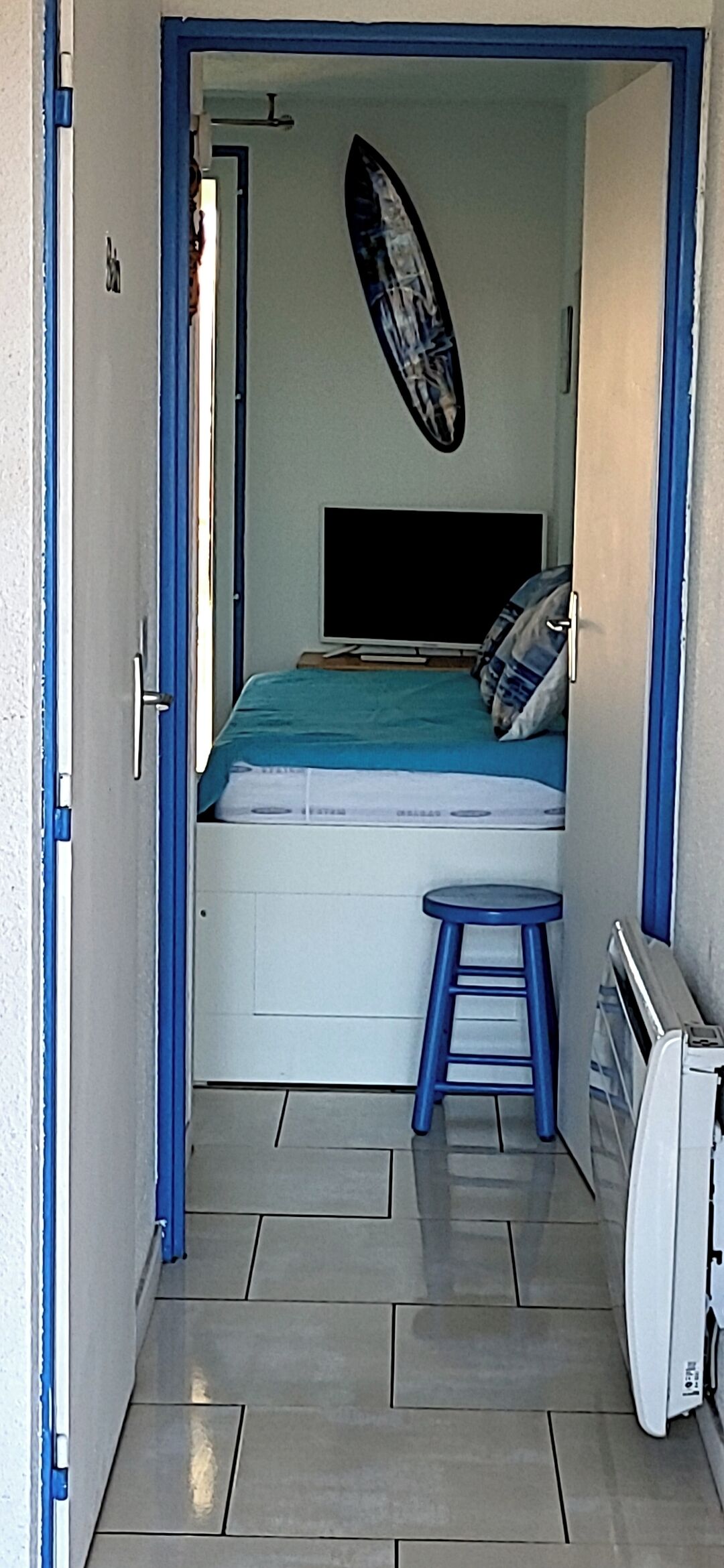 Ett litet rum med en säng, en blå pall, en tv och en surfbräda som hänger på väggen.