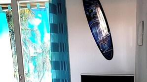 A bedroom with a bed, a TV, a surfboard decoration, and a view of palm trees through the door.