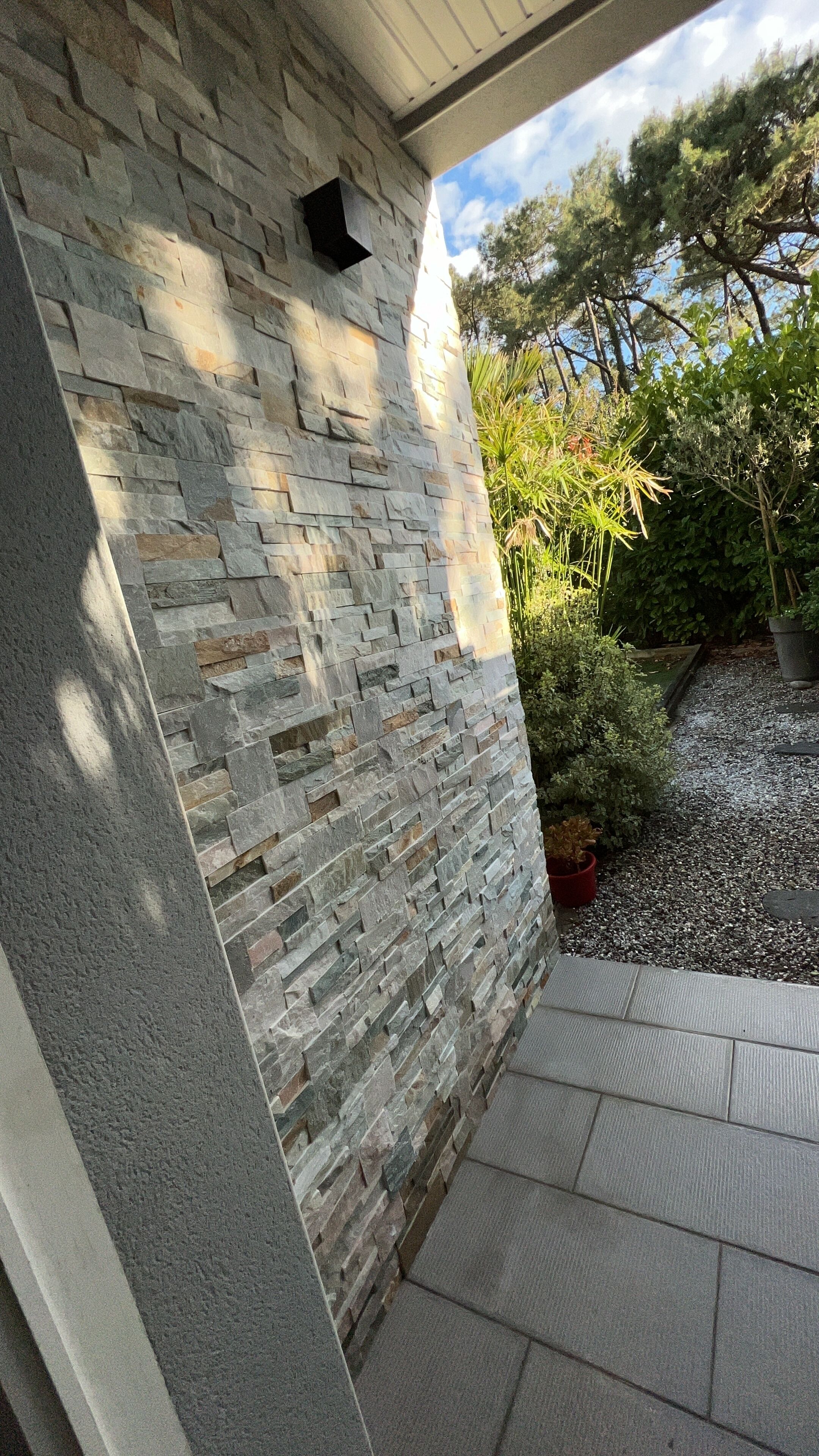Un mur de pierres avec une petite grille noire, un chemin de gravier et des plantes en pot.