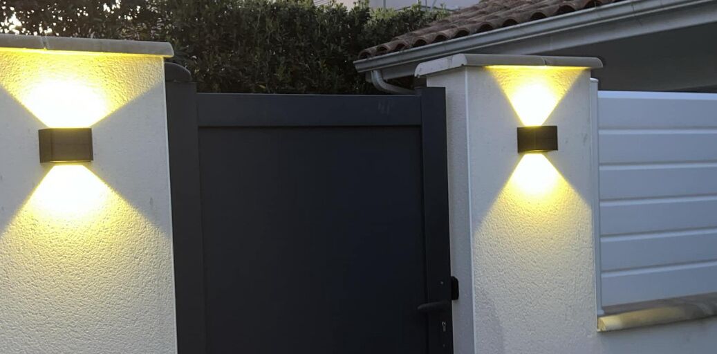 Una verja negra con dos luces empotradas a cada lado, una pared blanca y un tejado de tejas.