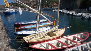 Uma marina com vários barcos pequenos atracados, um cais de pedra e um cenário de edifícios e falésias.
