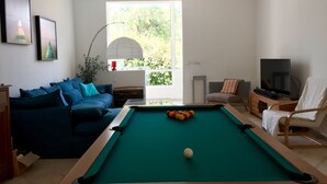 Een woonkamer met een pooltafel, een bank, een televisie en een raam met uitzicht op groen.