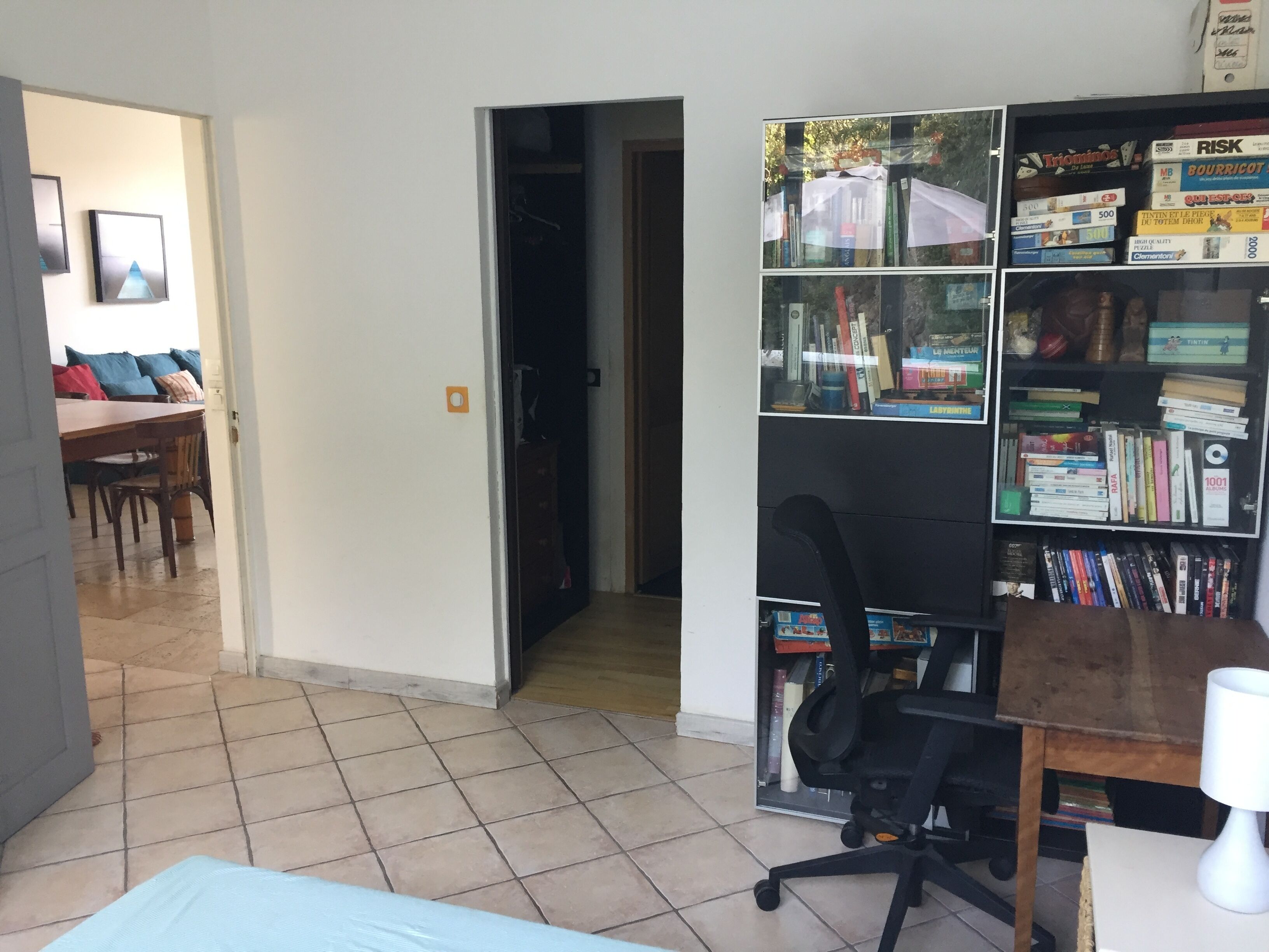 A room with a tiled floor, a desk with a chair, a bookshelf, and a bed.