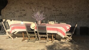 Un espacio de comedor al aire libre rústico, con un muro de ladrillo de fondo, una mesa con mantel a rayas, sillas de madera y una planta en maceta.