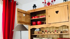 Una habitación con un armario de madera que contiene utensilios de cocina rojos, un florero con flores rojas y blancas sobre una mesa, y una cortina roja.