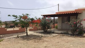 A residential area with a house featuring a tiled roof and a small porch. There are flowering plants and a paved pathway leading to the house. - Accommodation for 2 people, swimming pool, bikes in Morro île de Maio (Morro)